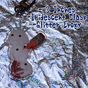 Puede incluir: Un dije de epoxi con purpurina en forma de mu&ntilde;eco de nieve con una botella roja, sujeto a un cierre met&aacute;lico. El dije mide aproximadamente 7,6 cm de largo. El texto "Frostcorpsclub" est&aacute; en la parte inferior de la imagen.