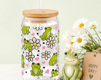 Puede incluir: Un vaso de vidrio transparente con tapa y pajita de bambú, decorado con patrones de ranas y flores verdes. Una figura de rana de cerámica y una etiqueta que dice "Hoppy Day!" están al lado.