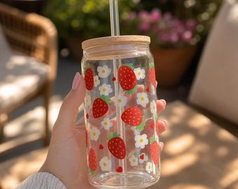 Puede incluir: Un vaso de vidrio transparente con tapa de bambú y pajita de vidrio. El vaso está decorado con fresas rojas y flores blancas. El vaso está sostenido en la mano de una persona.