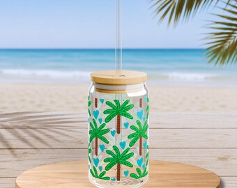 Puede incluir: Un vaso de cristal transparente con tapa de bambú y pajita de cristal, con un patrón repetitivo de palmeras verdes y pequeños corazones azul claro impresos en la superficie exterior del recipiente cilíndrico.
