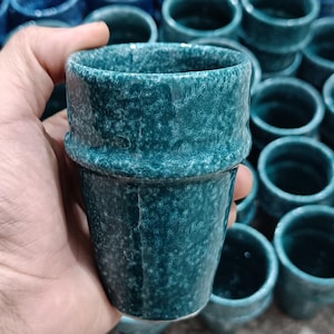 May include: A teal ceramic cup with a textured surface is held in a hand. The cup has a band around the middle. Many similar cups are in the background.