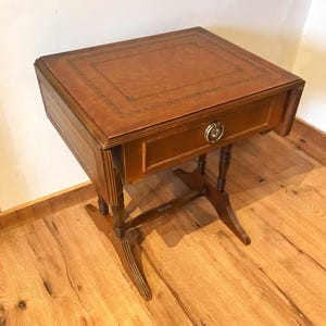 Puede incluir: Pequeña mesa auxiliar de madera antigua con tapa rectangular y un cajón. La tapa está cubierta de cuero marrón con un borde decorativo. La mesa tiene un acabado marrón oscuro y se apoya sobre cuatro patas.