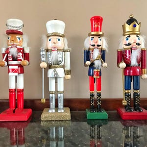 Op de afbeelding: Vier houten notenkrakers in verschillende outfits. De notenkrakers zijn geschilderd in rood, wit, blauw en goud. Elke figuur heeft een unieke hoed en gezichtskenmerken, staand op een rechthoekige basis. Een feestelijke vakantiedecoratie.