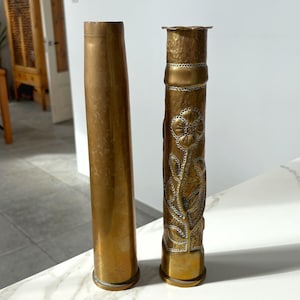 Peut inclure: Deux vases anciens en laiton. L'un est de forme cylindrique simple. L'autre est orné de motifs floraux et d'une surface texturée. Les deux vases ont une teinte dorée chaude et sont exposés sur une surface en marbre.