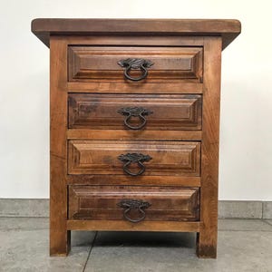 Puede incluir: Una mesita de noche de madera con cuatro cajones. La madera es de color marrón claro con veta visible. Cada cajón tiene un tirador decorativo de metal. La mesita es rectangular y tiene una tapa ligeramente saliente. Mide unos 60 cm de alto.