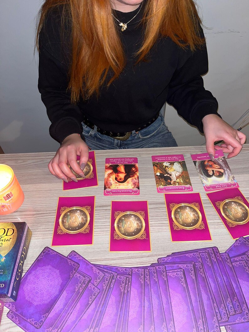 Op de afbeelding: Een tarotkaartopstelling met een pak paarse kaarten met sierlijke ontwerpen en een doos. Verschillende kaarten liggen op een houten oppervlak, met roze en gouden accenten. Een brandende kaars is zichtbaar.