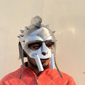 May include: A silver-colored gladiator helmet with a face mask and spiked details. The helmet has a crest on top and leather straps. The design is reminiscent of ancient Roman armor.