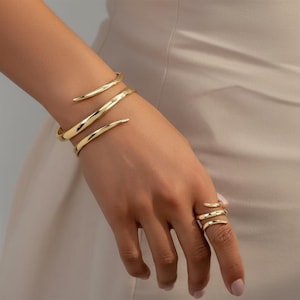 Puede incluir: Juego de joyas doradas con una pulsera y un anillo. La pulsera se envuelve alrededor de la muñeca con un diseño en espiral, y el anillo tiene un diseño en espiral similar. Las joyas se muestran en una mano.