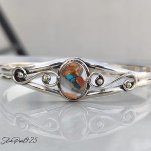 Könnte beinhalten: Ein silbernes Armreif-Armband mit einem ovalen, mehrfarbigen Stein in der Mitte. Der Stein weist Farbtöne von Orange, Blau und Weiß auf. Das Armband hat ein Doppelband-Design mit dekorativen Wirbeln und kleinen goldfarbenen Akzenten.