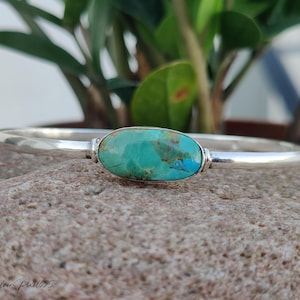 May include: A silver bangle bracelet with a large, oval turquoise stone at the center. The stone is a vibrant mix of blue and green hues. The bracelet is resting on a textured, gray stone surface, with green foliage in the background.