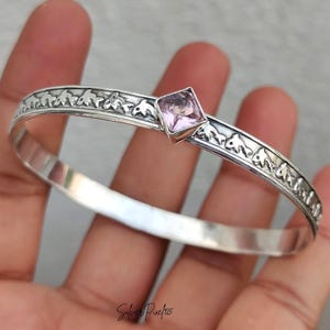 May include: A silver bangle bracelet featuring a repeating embossed pattern of small fish along the band. A single, square-cut, light purple gemstone is set prominently in the center. The text Silver Pixie925 is visible at the bottom of the image.