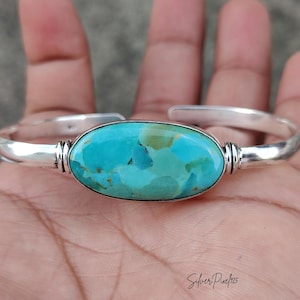 May include: A polished silver cuff bracelet featuring a large, oval-shaped turquoise stone with mottled blue and green hues. The metal band has a smooth, reflective finish with decorative grooved detailing on either side of the central gemstone.