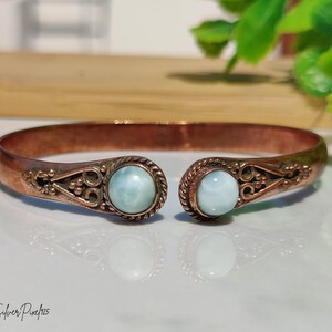 May include: A copper cuff bracelet featuring two round, light blue cabochon stones set at the open ends. The metal band displays intricate filigree scrollwork and twisted rope detailing surrounding each stone. The text Silver Pixe925 is visible.
