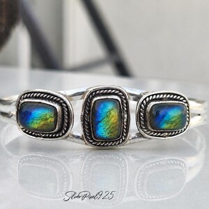 May include: A silver cuff bracelet featuring three rectangular labradorite stones. Each stone displays a vibrant play of color, including blues, greens, and yellows. The stones are set in a decorative, rope-like border.