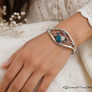 Könnte beinhalten: Ein silbernes Armreif-Armband mit einem ovalen Mittelstein aus Türkis, Rosa und Weiß. Das Armband hat ein filigranes Design und wird am Handgelenk getragen. Das Bild ist mit "Silver Pixel 925" signiert.