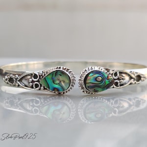 Peut inclure: Un bracelet manchette en argent avec deux incrustations de coquillages d'ormeau en forme de goutte d'eau. Les coquillages irisés affichent des teintes vertes, bleues et violettes. Le bracelet présente des détails argentés ornés.