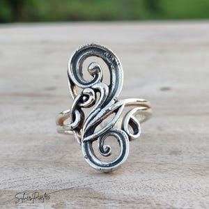 May include: A sterling silver ring featuring an elongated, openwork design with swirling, scroll-like patterns and oxidised detailing. The metal surface displays a polished finish with darkened recessed areas. The text Silver Pixie 925 is visible.