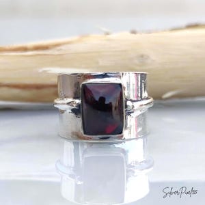 May include: A wide silver band ring featuring a rectangular, deep red garnet gemstone set in the center. The silver metal has a polished, reflective finish with visible texture and a prominent dark stone that catches the light.