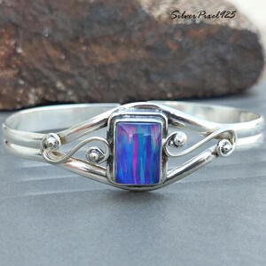 Peut inclure: Un bracelet jonc en argent avec une pierre centrale opale rectangulaire, irisée, bleue et rose. Le bracelet présente un design à double bande avec des volutes décoratives et de petits accents ronds. Le texte "Silver Pixel 925" est visible en arrière-plan.