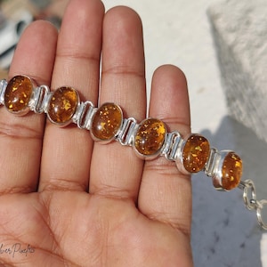 Peut inclure: Un bracelet en argent composé de sept pierres d'ambre ovales et translucides aux inclusions dorées. Les pierres sont serties dans des montures en argent poli et reliées par des maillons en argent, avec une chaînette d'extension. Texte : Silver Pixe/925.