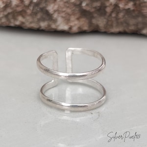May include: A silver ring featuring a double band design with an open gap at the front. The ring has a smooth, polished finish and a minimalist structure. The text Silver Pixel 925 is visible in the bottom right corner of the image.