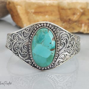 May include: A silver cuff bracelet featuring an oval turquoise stone in the centre. The metal surface is decorated with intricate scrollwork and a beaded border surrounding the stone. The text Silver Pixie 925 is visible in the bottom corner.