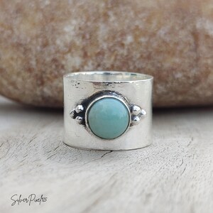 May include: A wide sterling silver band ring featuring a central round, smooth, light blue-green cabochon stone. The stone is flanked by small silver bead accents on both sides. The metal surface has a polished finish with subtle texture.
