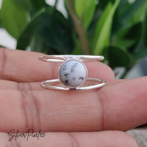 May include: A silver ring with a round, white stone featuring black speckles. The ring has a double-band design, with one band above and one below the stone. The ring is held in a hand, with green foliage in the background.