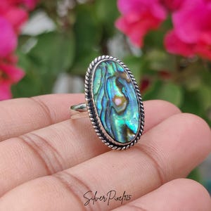 May include: Oval abalone shell ring with a silver band and a twisted rope design around the iridescent shell. The shell displays shades of blue, green, and pink. The ring is held in a hand against a blurred floral background.