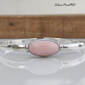 Peut inclure: Un bracelet en argent avec une pierre ovale rose clair au centre. Le bracelet a une texture martelée et reflète la lumière. Le texte "Silver Pixel 925" est visible en haut à droite.