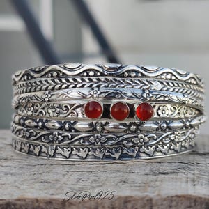 May include: A collection of silver bangles with detailed floral and wave designs. Three red gemstones are set in the central bangle. The bangles are displayed on a weathered wooden surface.