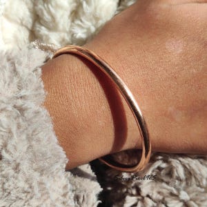 Puede incluir: Una pulsera de brazalete de color cobre, de aproximadamente 6,35 cm de diámetro, rodea una muñeca. La pulsera lisa y redondeada refleja la luz, creando reflejos. El fondo es una tela suave y texturizada de color blanquecino. El texto "Silver Pixel 925" es visible.