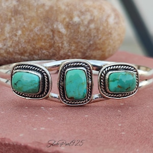 May include: A silver cuff bracelet featuring three rectangular turquoise stones. The stones are set in a decorative frame with a twisted rope design. The bracelet is displayed on a pink surface with a blurred background.