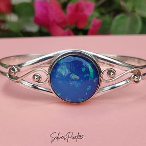 Puede incluir: Una pulsera de plata con una gran piedra de ópalo azul redonda en el centro. La pulsera tiene un diseño decorativo en espiral a cada lado de la piedra. El fondo es de color rosa suave.