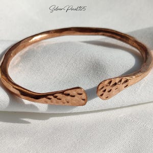 May include: A copper-colored open cuff bracelet with a hammered texture. The bracelet has an open design with rounded ends and a textured surface. The bracelet is displayed on a white fabric background. The text "Silver Pixel25" is in the upper left corner.