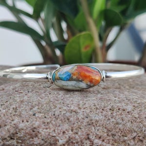 Könnte beinhalten: Ein silbernes Armreif-Armband mit einem ovalen, mehrfarbigen Stein-Mittelstück. Der Stein weist Farbtöne von Orange, Blau und Weiß auf. Das Armband wird auf einer strukturierten, hellbraunen Oberfläche präsentiert.