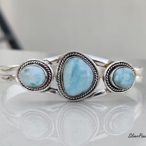 May include: A silver bracelet featuring three light blue larimar stones. The central stone is a unique, irregular shape, flanked by two oval stones. Each stone is set in a decorative, rope-like border.