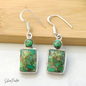 May include: A pair of silver drop earrings featuring a small round turquoise stone above a larger rectangular turquoise stone. Each stone displays a vibrant green hue with intricate copper-coloured veining. The earrings hang from silver hooks.