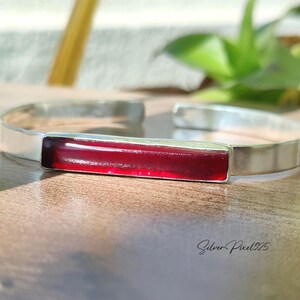 May include: A silver cuff bracelet featuring a rectangular, deep red gemstone. The bracelet has a polished finish and a minimalist design. The jewellery is displayed on a wooden surface, suitable for everyday wear.