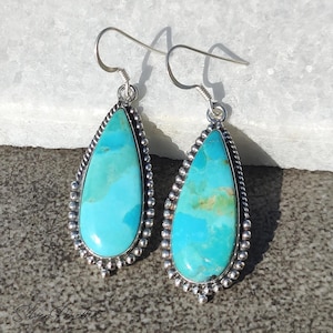 May include: A pair of turquoise teardrop earrings with a silver beaded border and silver hooks. The turquoise stones have natural variations in color, ranging from light to medium blue. The earrings are displayed on a light-colored surface.