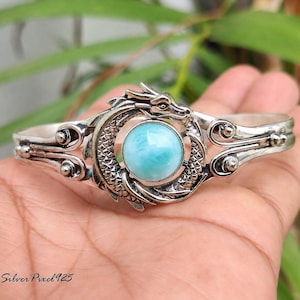 Könnte beinhalten: Ein silberfarbenes Armband mit einem Drachendesign. Das Armband hat einen runden, hellblauen Edelstein in der Mitte. Der Drachenkopf befindet sich oben, der Schwanz umschließt den Edelstein. Das Armband ist unten offen.