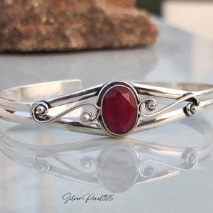 Peut inclure: Un bracelet manchette en argent orné d'un rubis ovale et facetté. Le bracelet présente un design à double bande avec des volutes décoratives et de petits accents dorés. Le métal poli reflète la lumière, rehaussant la couleur rouge profond du rubis. C'est un bijou.