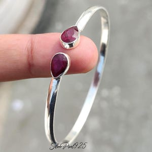 May include: A silver cuff bracelet with two teardrop-shaped, deep red gemstones. The gemstones are set in a polished silver band. The bracelet is shown against a blurred background.