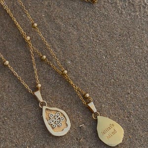 May include: Two gold necklaces with teardrop pendants. One pendant has an intricate design, the other is engraved with "Jelitzl's Mind." The necklaces lie on a textured, sandy surface.