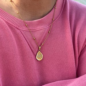 May include: A gold necklace with a teardrop pendant and an ornate design is worn over a pink sweatshirt. The necklace has a fine chain with small, round beads. The sweatshirt is a crew neck style.