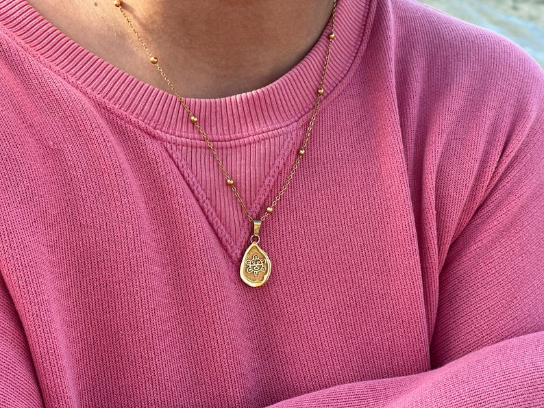 May include: A gold necklace with a teardrop pendant and an ornate design is worn over a pink sweatshirt. The necklace has a fine chain with small, round beads. The sweatshirt is a crew neck style.