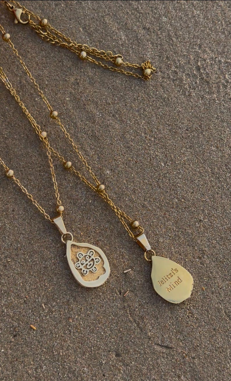 May include: Two gold necklaces with teardrop pendants. One pendant has an intricate design, the other is engraved with "Jelitzl's Mind." The necklaces lie on a textured, sandy surface.
