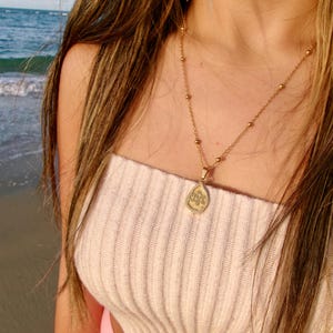 May include: A gold necklace with a pendant featuring an engraved design. The necklace is worn over a cream-coloured, ribbed tube top. The background shows a beach with ocean waves.