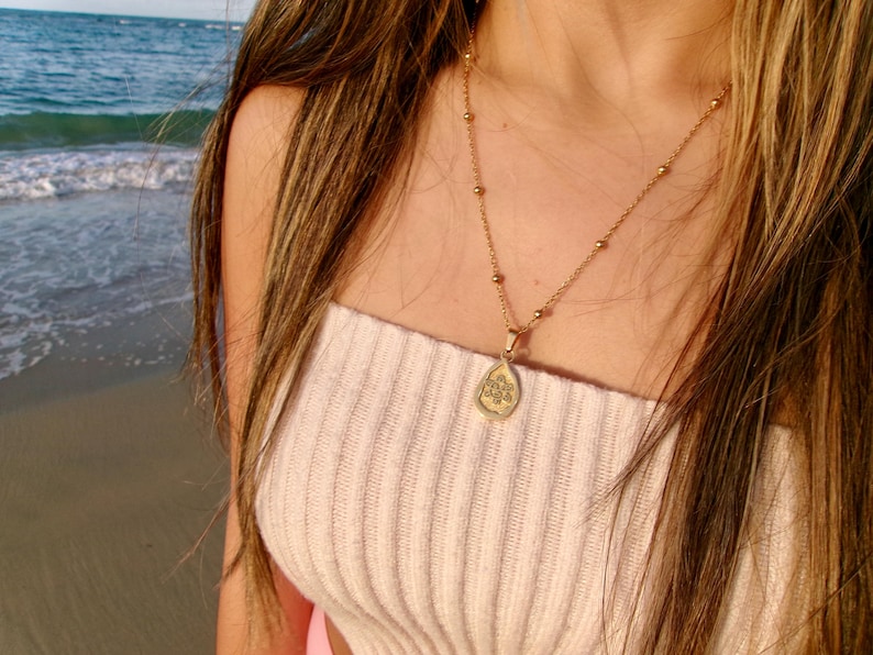 May include: A gold necklace with a pendant featuring an engraved design. The necklace is worn over a cream-coloured, ribbed tube top. The background shows a beach with ocean waves.