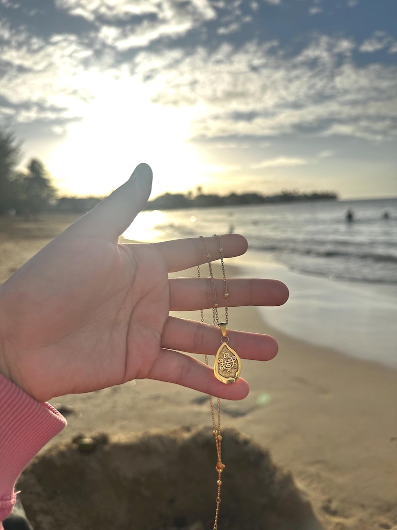 Puede incluir: Un collar con colgante dorado con un dije en forma de lágrima. El dije presenta un diseño intrincado. El collar se sostiene contra un fondo de playa con el océano y el cielo al fondo.